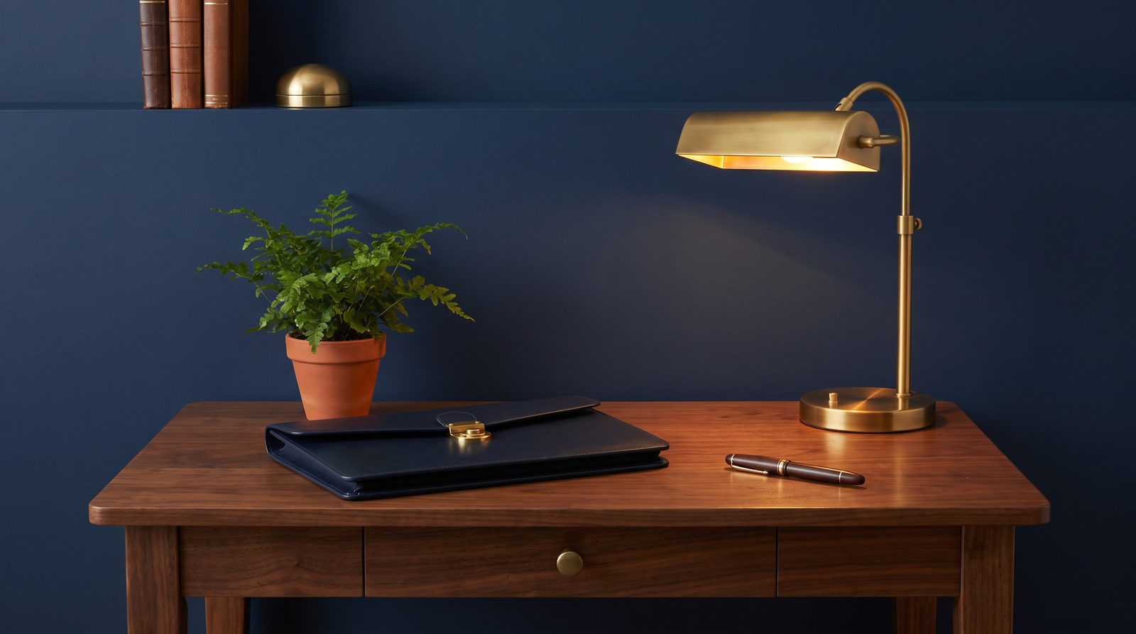 A navy leather portfolio, brass desk lamp and potted fern on a tidy walnut desk
